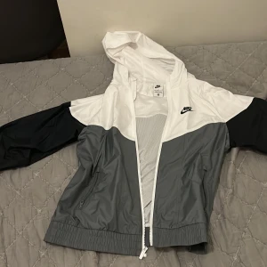Nike vindjacka med huva i grått och vitt - Nike vindjacka med huva i stilren färgblockad design. Jackan har vita och grå paneler med svarta ärmar, hel dragkedja framtill och två sidofickor med dragkedja. Perfekt för att hålla vinden borta och samtidigt se fräsch ut.