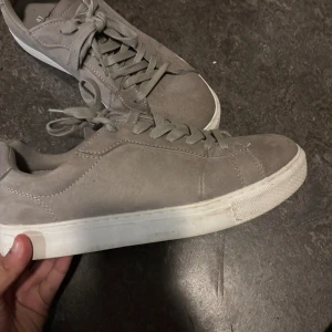 Grå mockasneakers med vit sula - Snygga grå sneakers i mocka med klassisk design och vit platt sula. Skorna har rund tå, snörning och diskreta sömmar som ger en stilren look. Perfekt för dig som gillar enkel och clean stil. Storlek 41. Använda 2 gånger. 