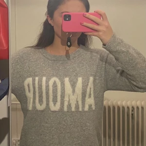 Grå stickad tröja med vit text AMOUR - Mysig grå stickad tröja med stor vit fluffig text AMOUR över bröstet. Rund halsringning och långa ärmar. Perfekt för dig som gillar chill och enkel stil med en twist. Passar dig som vill ha en skön och snygg tröja till höst och vinter.