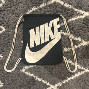 Svart Nike gympåse  - Svart gympåse från Nike med stor vit logga framtill. Väskan har vita snören och en praktisk dragkedjeficka på sidan. Tillverkad i lätt syntetmaterial, perfekt för att bära träningskläder eller skor. Enkel och stilren design som funkar till det mesta.