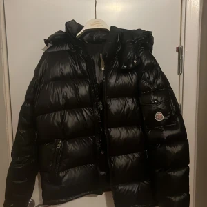 Svart Moncler pufferjacka med huva - Svart pufferjacka från Moncler med glansig finish och huva. Jackan har dragkedja framtill, sidofickor med dragkedja och Moncler-logga på ärmen. Insidan har ett unikt serietryck och klassisk Moncler-etikett. Perfekt för kalla dagar. Använts några gånger men är som ny! Kontakta för frågor osv.
