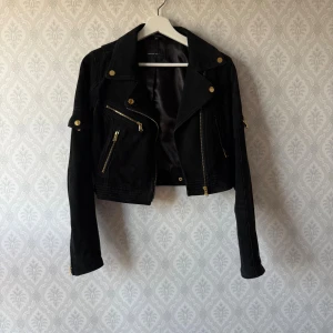 Svart mocka skinnjacka Rock'n Blue - Snygg svart jacka i mocka från Rock'n Blue med guldiga detaljer och dragkedjor. Croppad modell med bred krage och flera fickor framtill. Jackan har lång ärm. Perfekt för dig som vill ha en statement piece i garderoben. Knappt använd. Rökfritt hem.