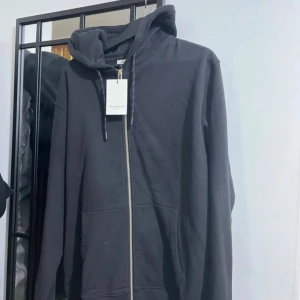 Svart hoodie med dragkedja och huva - Svart hoodie med hel dragkedja framtill och stor huva. Tröjan har snörning vid halsen och två rymliga fickor på magen. Perfekt för chill dagar och enkel att matcha med jeans eller joggers.