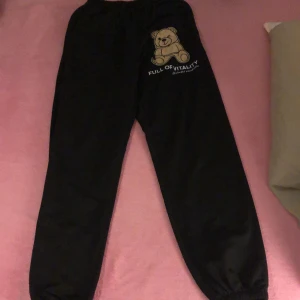 Svarta mjukisbyxor/sweatpants - Svarta mjukisbyxor med elastisk midja och muddar vid bensluten. På ena benet finns ett beige nalletryck med texten 'FULL OF VITALITY Unlimited possibilities'. Sköna och chill byxor, perfekta för att hänga hemma eller dra till gymmet.