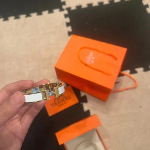  vit och guld armband - Säljer ett stilrent  armband i vit och guld med klassisk H-detalj framtill. Armbandet är i metall med vit emalj och har en elegant, rundad form. Kommer med original ask, påse och certifikat. Perfekt för dig som gillar lyxiga accessoarer.