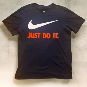 Nike t-shirt med Just Do It tryck - Mörkblå t-shirt från Nike med klassisk vit swoosh och stort rött 'JUST DO IT.' tryck på bröstet. Rund hals och korta ärmar. Tillverkad i mjuk bomull som känns skön mot huden. Perfekt för dig som gillar sportig och clean stil. Rökfritt hus!