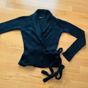 Svart omlottkofta från Gina Tricot - Mysig svart stickad omlottkofta från Gina Tricot med långa ärmar och bred krage. Koftan knyts i midjan med ett matchande band för en snygg och feminin siluett. Perfekt att slänga på sig när det är lite kyligare ute.