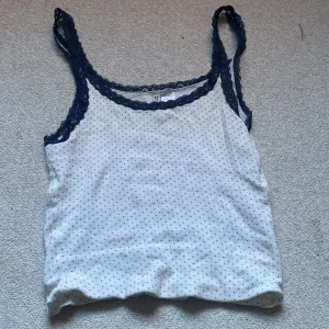 Vit prickig linnetopp med spets från DIVIDED - Säljer en söt vit linnetopp från DIVIDED med små mörkblå prickar och mörkblå spets runt halsringning och axelband. Har varit SLUTSÅLD.  Toppen är ärmlös och har en mjuk, ribbad känsla. Perfekt för att ge din outfit en chill och feminin vibe.