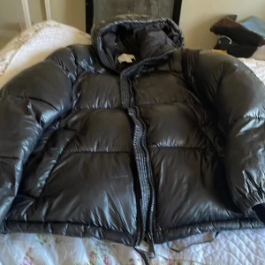 Svart pufferjacka från H&M, S - Svart pufferjacka från H&M i storlek S. Jackan har en glansig yta, huva och dragkedja framtill. Tillverkad i 100% polyester med elastiska muddar vid ärmslut. Perfekt för kalla dagar när du vill ha en stilren och varm look.Passar till dom som har storlek M med då den är stor i storlek.