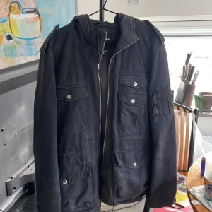 Svart parkajacka Calvin Klein Jeans - Svart parkajacka från Calvin Klein Jeans med huva, flera fickor med tryckknappar och dragkedja framtill. Jackan har en clean look med snygga detaljer på axlarna