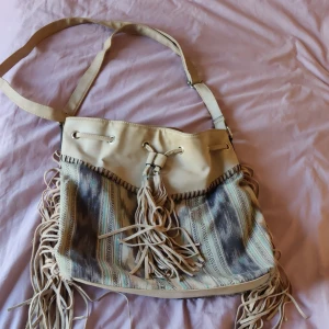 Beige axelväska med fransar och mönster - Säljer en beige axelväska med coola fransar på sidorna och snörning upptill. Väskan har ett etniskt mönster i blått, grått och beige, samt en dragkedja på baksidan. Insidan är fodrad i korallfärgat tyg och har flera fack. Perfekt för dig som gillar boho-stil!