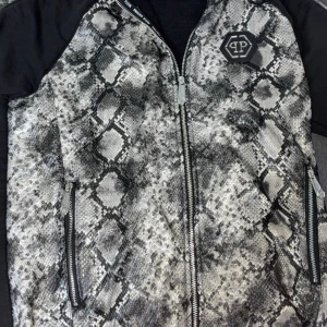 Philipp Plein bomberjacka med panda - Säljer en unik bomberjacka från Philipp Plein med svart- och grått ormmönster på framsidan, coolt panda-motiv med paljetter på ryggen och detaljerade dragkedjor. Jackan har svarta ärmar och flera snygga logopatchar. Perfekt för dig som vill sticka ut.