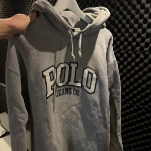 Grå hoodie Polo Jeans Co. Ralph Lauren - Snygg grå hoodie från Polo Jeans Co. Ralph Lauren med stor broderad logga på bröstet. Tröjan har huva med snörning och långa ärmar. Tillverkad i mjuk bomull och polyester, perfekt för chill dagar. Klassisk och enkel stil som funkar året runt.