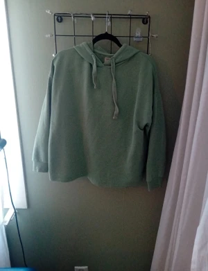 Grön hoodie med huva och snörning - Snygg grön hoodie med huva och snörning framtill. Tröjan har en loose passform och långa ärmar, perfekt för chill dagar. Materialet känns mjukt och skönt mot huden. Enkel design utan mönster eller logga, passar dig som gillar stilrena plagg.