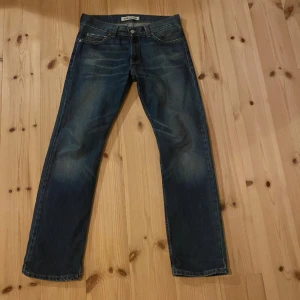 Levi's 506 Standard jeans blå - Klassiska Levi's 506 Standard jeans i blå denim med snyggt slitna detaljer och raka ben. Jeansen har fem fickor, orange sömmar och en autentisk läderpatch bak i midjan. Perfekta för en avslappnad och tidlös stil.