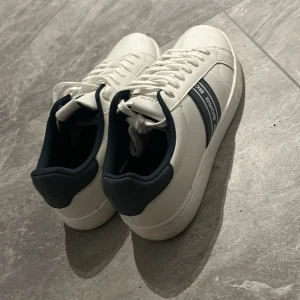 Vita sneakers från Björn Borg - (Använda en gång!) Snygga vita sneakers från Björn Borg med marinblå detaljer och logga på sidan. Skorna har snörning, rund tå och platt sula. Tillverkade i skinnimitation med mjukt foder och klassisk siluett. Perfekta för dig som gillar stilrena och sportiga skor. Pris går givetvis att diskutera! 