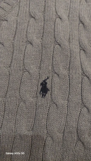 Grå kabelstickad tröja Polo Ralph Lauren - Kabelstickad tröja i ljusgrått från Polo Ralph Lauren, storlek XL. Klassisk rund hals och långärmad modell med broderad marinblå logga på bröstet. Mjuk och skön känsla, perfekt för kyliga dagar. Ribbstickade muddar vid ärmslut och nederkant.