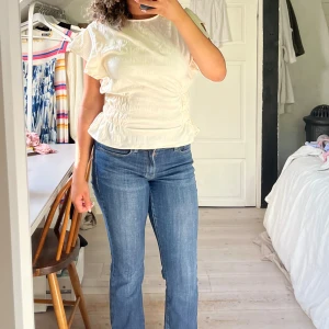 Vit topp med rynkdetaljer och volanger - Säljer en vit topp med rynkdetaljer och volanger. Tröjan är stretchig vid midjan. Råkade köpa två och har därför är tröjan inte använd.