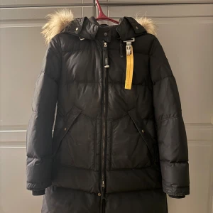 Svart Parajumpers lång dunjacka - Säljer en svart lång dunjacka från Parajumpers med fluffig päls på huvan och den klassiska gula remmen vid halsen. Jackan har två stora fickor fram, dragkedja och knappar, samt en varm och quiltad design som är perfekt för kalla dagar. Nypris 8000:-