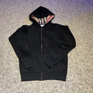 Svart Burberry hoodiejacka med dragkedja - Svart hoodiejacka från Burberry med klassisk rutig huva i beige, rött och svart. Jackan har dragkedja i guld, snörning i huvan och känguruficka framtill. Perfekt för dig som vill ha en stilren och lyxig streetwear-look.