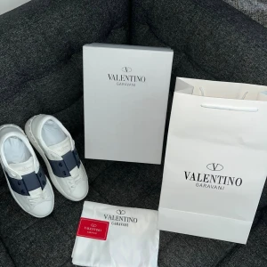 Valentino Garavani Open skor - Säljer ett par Valentino Garavani Open sneakers i vitt skinn med bred marinblå rem över vristen. Klassisk rund tå, platt sula med mönstrad undersida och snygga snörningar. Lyxig och stilren design, perfekt för dig som gillar exklusiva sneakers.
