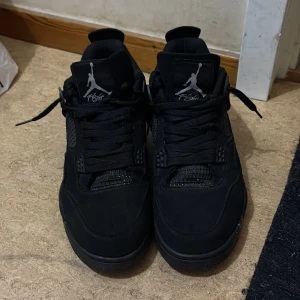 Svarta Nike Air Jordan 4 sneakers - Säljer ett par svarta Nike Air Jordan 4 sneakers med klassisk Jumpman-logga på plösen och hälen. Skorna har snörning, platt sula och meshdetaljer på sidorna. Materialet är en mix av skinn och syntet, vilket ger en snygg och sportig look. Perfekt för dig som gillar streetwear.
