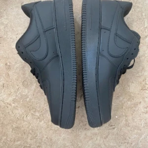 Svarta Nike Air Force 1 sneakers - Säljer ett par helsvarta Nike Air Force 1 sneakers med klassisk låg siluett och rund tå. Skorna har snörning och är tillverkade i skinn med en platt, robust sula. Swoosh-loggan syns på sidorna och Air-detalj på sulan. Perfekt för dig som gillar stilrena och tidlösa sneakers.