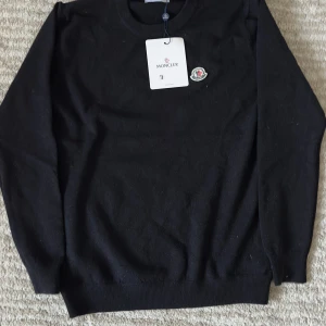 Svart Moncler tröja - Snygg svart stickad tröja från Moncler med rund halsringning och klassisk logga på bröstet. Tröjan har långa ärmar och ribbade muddar för en clean look. Perfekt för dig som gillar stilrena och exklusiva plagg.