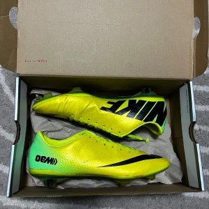 Nike Mercurial fotbollsskor gul/svart - Nike Mercurial fotbollsskor i en riktigt fet gul färg med svarta detaljer och logga. Skorna har snörning, platt sula med dobbar och en rund tå. Ovandelen är gjord i syntetmaterial med en texturerad yta för bättre bollkontroll. Perfekt för dig som vill sticka ut på planen.