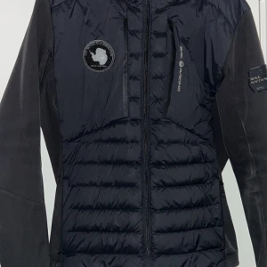 Svart Sail Racing jacka - Säljer en svart jacka från Sail Racing med huva och dragkedja. Jackan har ett broderat märke på bröstet och är quiltad för extra värme. Perfekt för kyligare dagar.