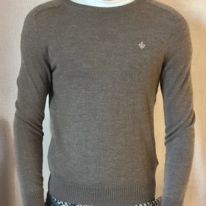 Brun merinoullströja från Morris XS - Snygg brun stickad tröja från Morris i 100% merinoull. Klassisk rund halsringning, diskret logga broderad på bröstet och ribbade muddar vid ärmslut och nederkant. Perfekt för lager-på-lager och stilren look. Tröjan är nästan helt oanvänd eftersom den inte passar