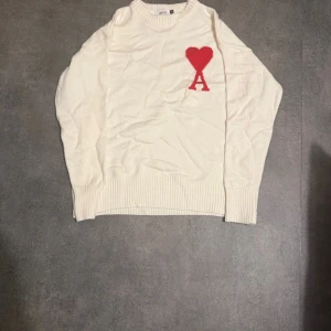 Vit stickad tröja från Ami med rött hjärta - Vit stickad tröja från Ami med rund halsringning och ribbade muddar. På bröstet finns ett stort rött hjärta med bokstaven A. Tröjan har en clean look och är perfekt för dig som gillar stilrena detaljer.