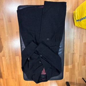 Svart kappa från Lindex med bälte - Säljer en svart kappa från Lindex i storlek 36. Jackan har ett snyggt bälte i midjan, knappar framtill och ett mönstrat rött innerfoder. Materialet är grovstickat och ger en cool struktur. Perfekt för dig som gillar klassisk stil med en twist.