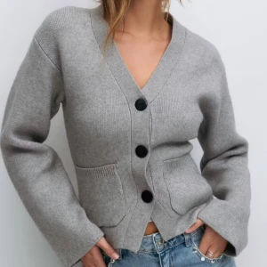 Grå kofta från Gina Tricot - Grå kofta med v-ringning och stora svarta knappar från Gina Tricot. Modellen har två fickor framtill och är stickad i mjukt material. Långärmad och med normal passform, perfekt att slänga över en t-shirt eller linne. Enkel och clean look.