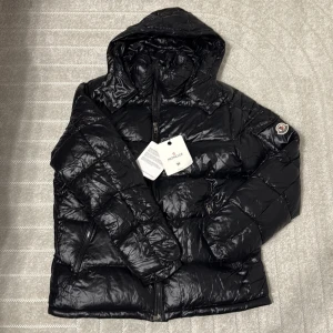 Svart Moncler pufferjacka med huva - Svart pufferjacka från Moncler med glansig finish, huva och dragkedja framtill. Jackan har Moncler-logga på ärmen och coola detaljer som innerfoder med serietidningsmotiv. Perfekt för dig som vill ha en varm och snygg jacka med streetkänsla.