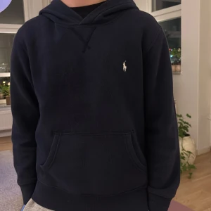 Mörkblå hoodie från Polo Ralph Lauren - Snygg mörkblå hoodie från Polo Ralph Lauren med klassisk logga broderad på bröstet. Tröjan har huva, känguruficka och långa ärmar. Perfekt för chill dagar och enkel att matcha med jeans eller mjukisbyxor.