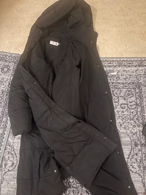 Svart lång pufferjacka med luva - Säljer en svart lång pufferjacka från Nelly med luva och tryckknappar med dragkedja längs hela framsidan. Jackan har quiltad design och är perfekt för kalla dagar. Materialet är syntet och jackan har långa ärmar. Enkel och stilren look som funkar till det mesta.