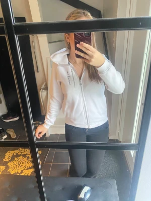 Vit hoodie från Sail racing med dragkedja - Säljer en fräsch vit hoodie från Sail racing  med hel dragkedja framtill och en extra dragkedja vid bröstet. Tröjan har huva och långa ärmar, perfekt för chill dagar. Svea-loggan är broderad på bröstet. Skön och enkel stil som funkar till det mesta. Storleken är egentligen 160 men passar mig