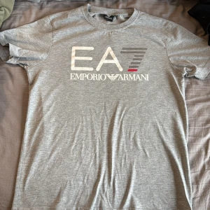Grå EA7 Emporio Armani t-shirt - Snygg grå t-shirt från EA7 Emporio Armani med stort logotryck framtill. Klassisk rund halsringning och korta ärmar. Tillverkad i mjuk bomull för en skön känsla. Perfekt för dig som gillar stilrena och exklusiva plagg.