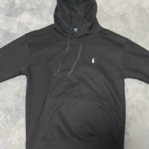 Svart hoodie från Polo Ralph Lauren - Svart hoodie från Polo Ralph Lauren med klassisk logga broderad i vitt på bröstet. Tröjan har huva med snörning, stor magficka och långa ärmar. Perfekt för chill dagar och enkel att matcha med jeans eller joggers.