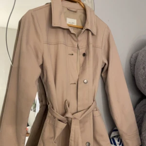 Beige trenchcoat från Cellbes - Snygg beige trenchcoat från Cellbes med klassisk krage, knappar och knytskärp i midjan. Jackan har långa ärmar och praktiska fickor framtill. Perfekt för dig som gillar stilrena och tidlösa plagg med enkel design 💗