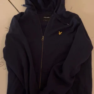 Mörkblå hoodiejacka från Lyle & Scott - Snygg mörkblå hoodiejacka från Lyle & Scott med gul broderad logga på bröstet. Jackan har huva, dragkedja framtill och är långärmad. Perfekt för chill dagar och enkel att matcha med jeans eller joggers.