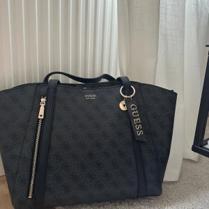 Svart väska från Guess - Helt oanvänd svart väska från Guess med subtilt mönster och guldfärgade detaljer. Väskan är helt ny då jag fick den i födelsedagspresent men fick dubbletter. PRIS KAN DISKUTERAS!!Väskan har en stor dragkedja framtill, två handtag och en matchande innerficka med dragkedja. Guess-logga i metall och snygg tag med logotyp. Perfekt storlek för allt du behöver.