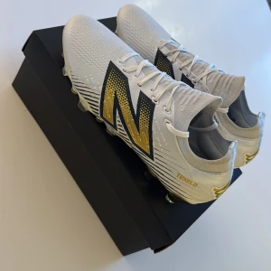 New Balance Tekela vit/guld fotbollsskor - Säljer ett par New Balance Tekela fotbollsskor i vit med stora guld- och svarta detaljer. Skorna har snörning, platt sula med guldiga dobbar och en strömlinjeformad design. Materialet är syntet och mesh för lätthet och komfort på planen.