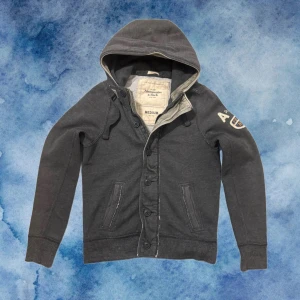 Mörkgrå Vintage Abercrombie & Fitch Cardigan  - Mörkgrå Caridgan från Abercrombie & Fitch med broderad logga och emblem på ärmen. Tröjan har knappar framtill, huva och två fickor. Tröjan har dessutom knappar + dragkedja att stänga med, perfekt till vintern. Längd ≈ 65 cm , Bred Armhåla - Amrhåla ≈ 54cm. Pris kan diskuteras!