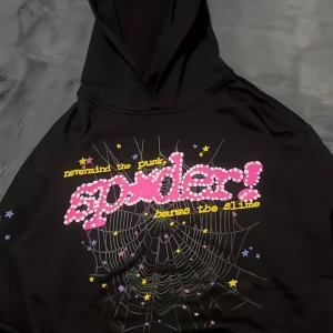 Svart hoodie med spindelnätsprint - Cool svart hoodie med stor färgglad print på bröstet med texten 'spider!' i rosa och vitt, samt stjärnor och spindelnät i gult, lila och rosa. Tröjan har huva och passar dig som gillar grafiska tryck och streetstyle.