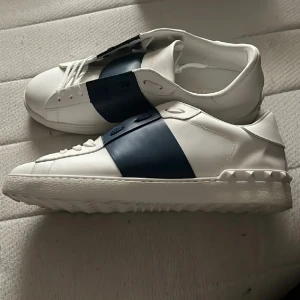 Valentino Garavani vita sneakers - Snygga vita sneakers från Valentino Garavani med bred marinblå läderdetalj över vristen och ikoniska nitar på sulan. Skorna har snörning och är tillverkade i skinn, vilket ger en lyxig känsla. Perfekta för dig som vill sticka ut med stilrena designerdojor. Nypris 3499kr dom passar även storlek 43