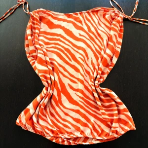 Orange zebramönstrad klänning Michael Kors - Snygg kortklänning från Michael Kors i orange och vitt zebramönster. Klänningen har tunna axelband med knyt och silverfärgade MK-detaljer. Tillverkad i ett lätt och luftigt material som passar perfekt för varma dagar. Modellen är loose och har en avslappnad passform.