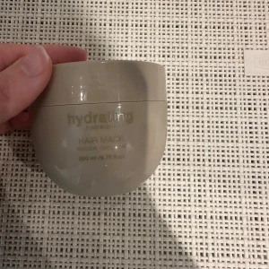 Miqura Hydrating Hair Mask 200ml - Beige hårinpackning från Miqura i en rund plastburk på 200 ml. Hydrating Hair Mask ger intensiv återfuktning och vård till håret. Snygg minimalistisk design med guldig text och lock. Perfekt för dig som vill boosta håret med extra fukt.