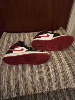 Nike Air Jordan 1 High röd/svart/vit - Nike Air Jordan 1 High sneakers i klassisk färgkombination med rött, svart och vitt. Storlek: 40.5. Skorna har högt skaft, svart snörning och perforerad tå för extra ventilation. Ikonisk röd swoosh på sidan och mjuk vadderad krage. Materialet är skinn och syntet.
